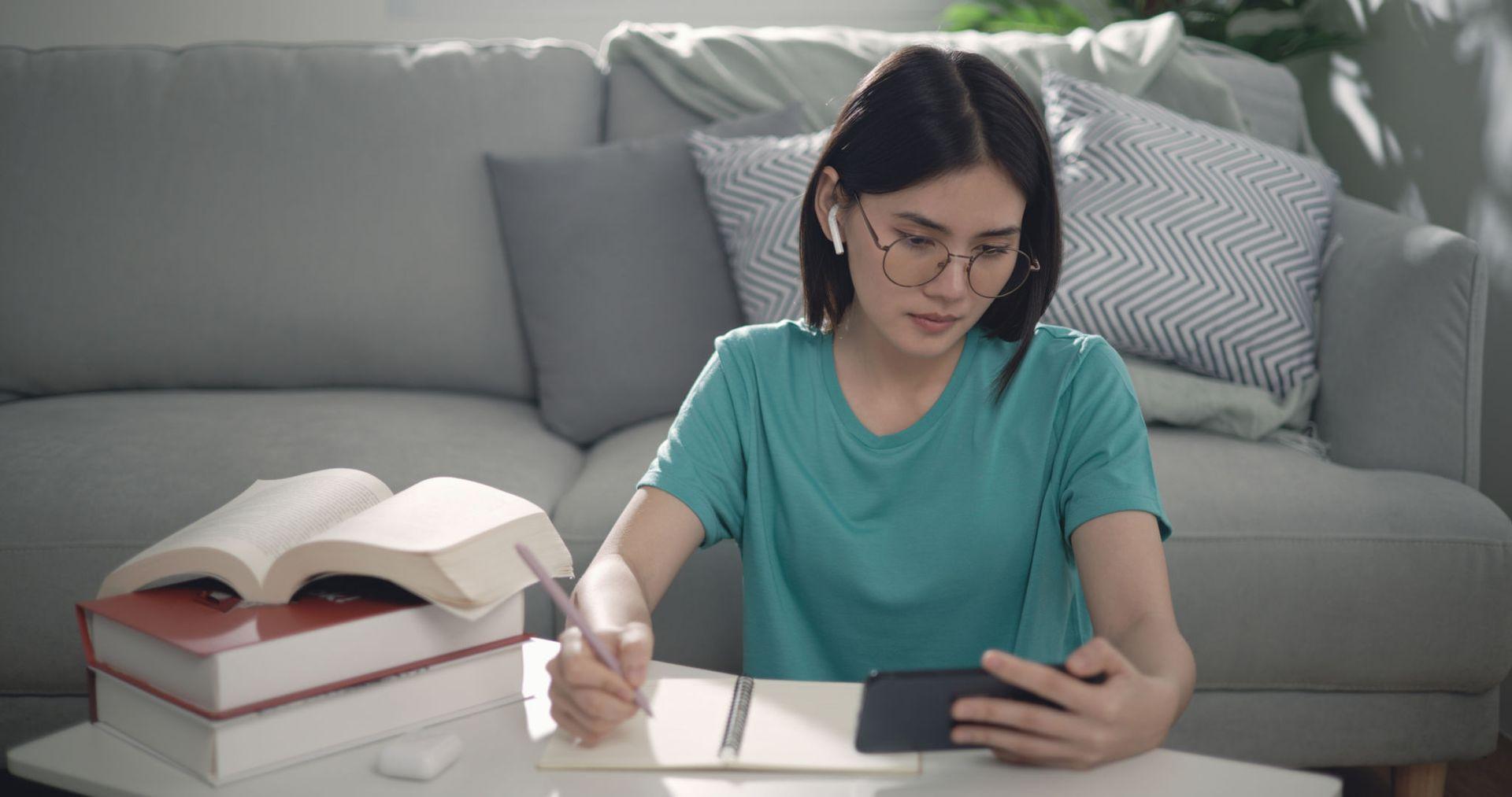 Person with glasses studying with books, writing in a notebook while looking at a smartphone.